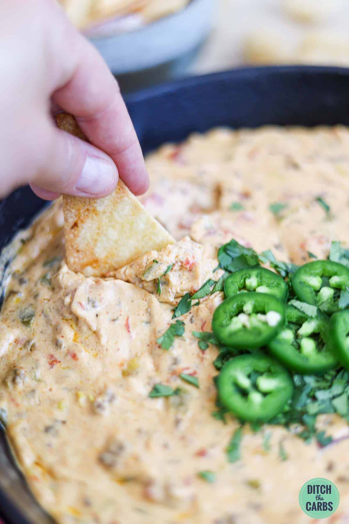 A hand kipping a keto tortilla chip into a skillet full of meaty keto queso dip.