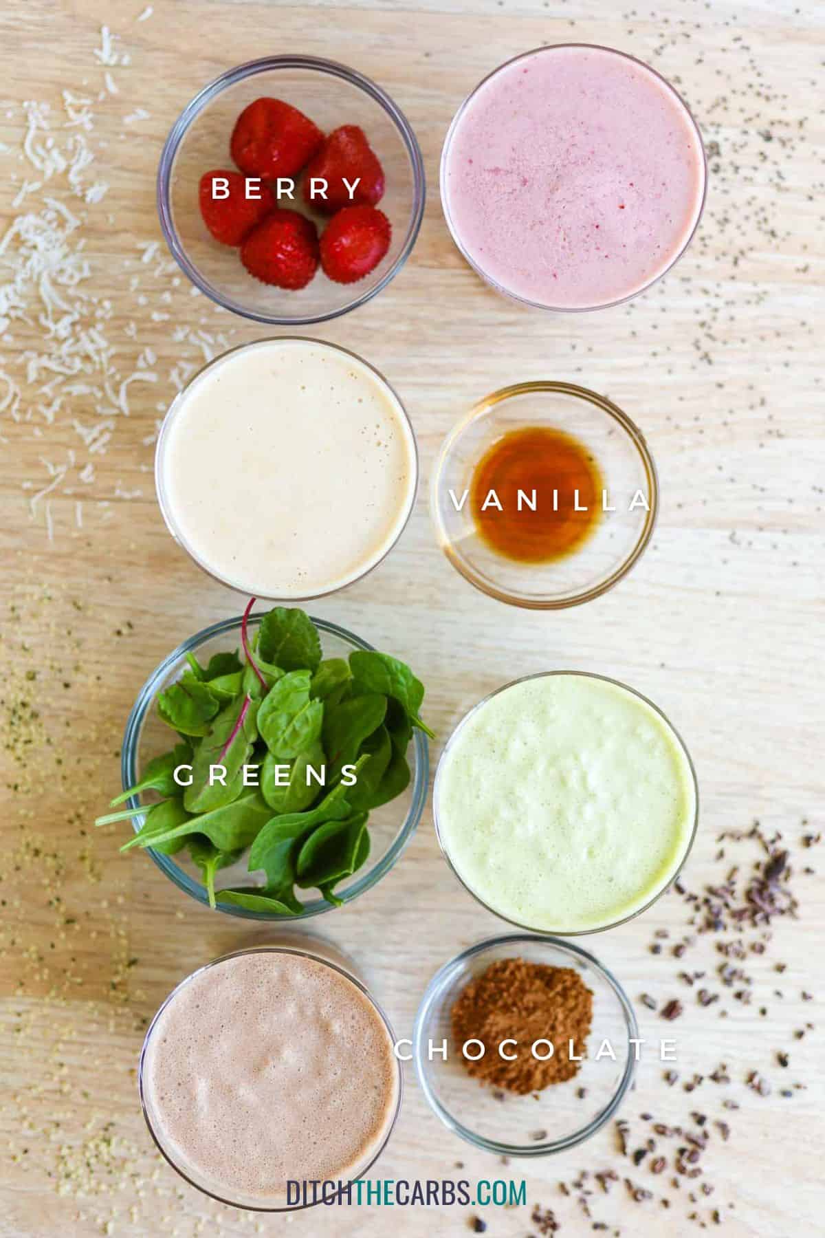 4 variations of low-carb protein shakes in glass cups with a bowl of the variation ingredients next to it. The berry flavor has strawberries. The Vanilla flavor has vanilla extract next to it. The green variation has a handful of baby spinach, and the chocolate version has a tablespoon of cocoa powdered next to it.