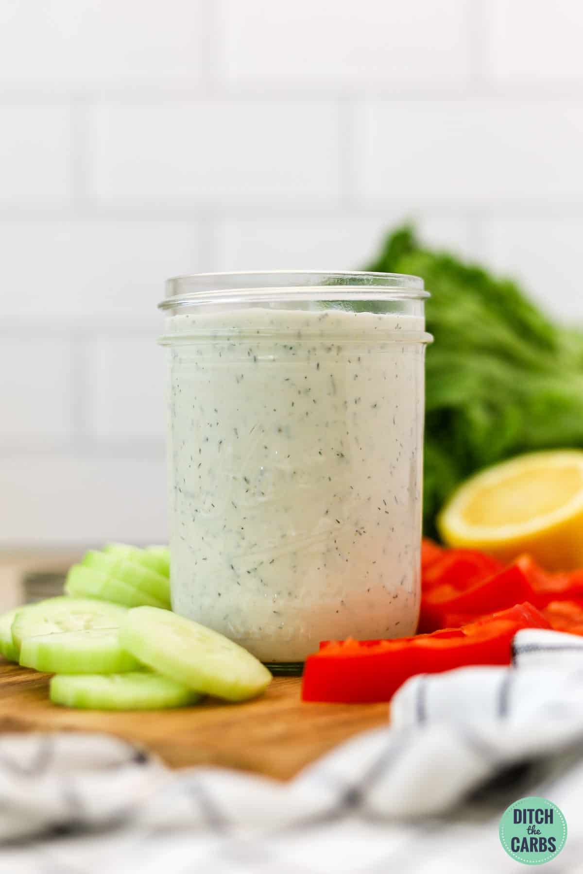 A jar of keto ranch dressing on a wooden tray with sliced raw vegetables around it.
