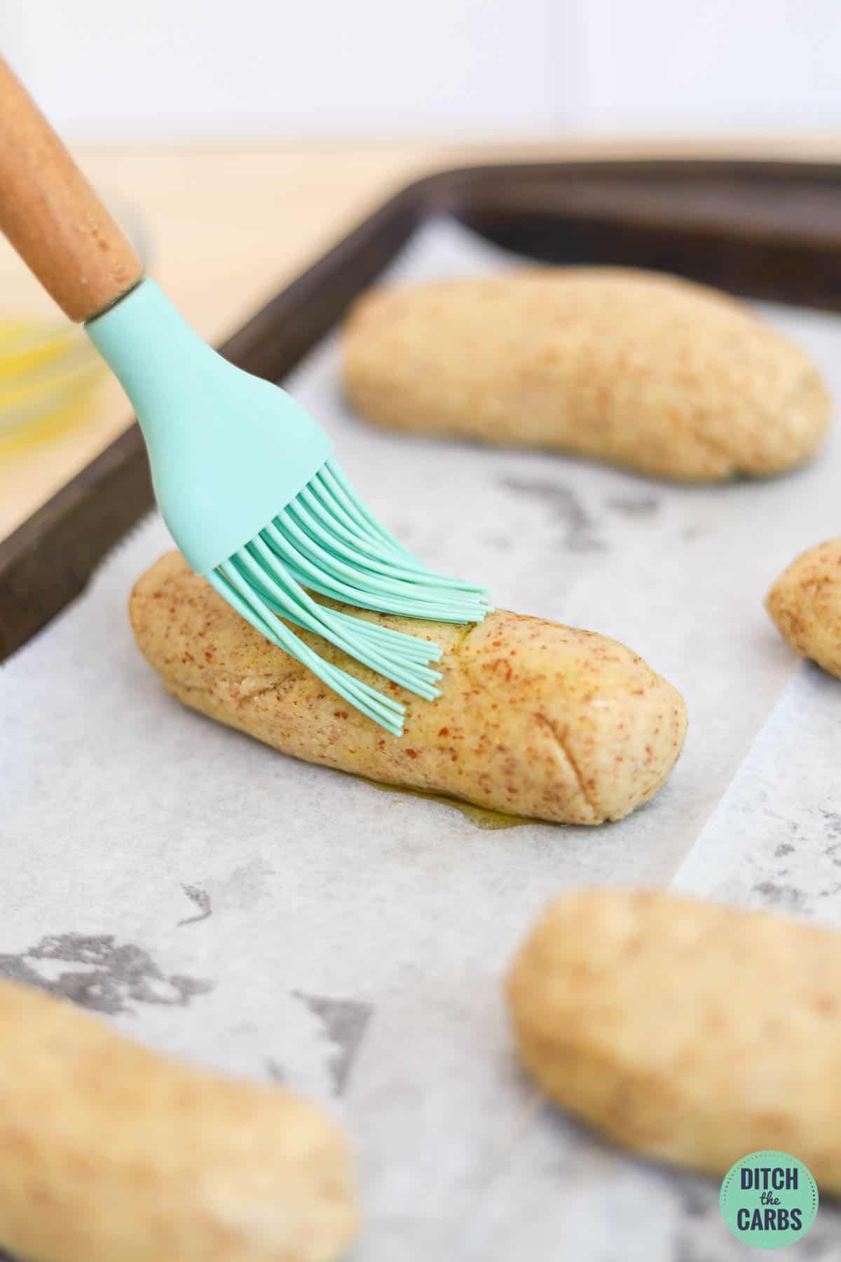 A basting brush is spread olive oil over a shaped keto hot dog bun on a lined baking tray before baking.
