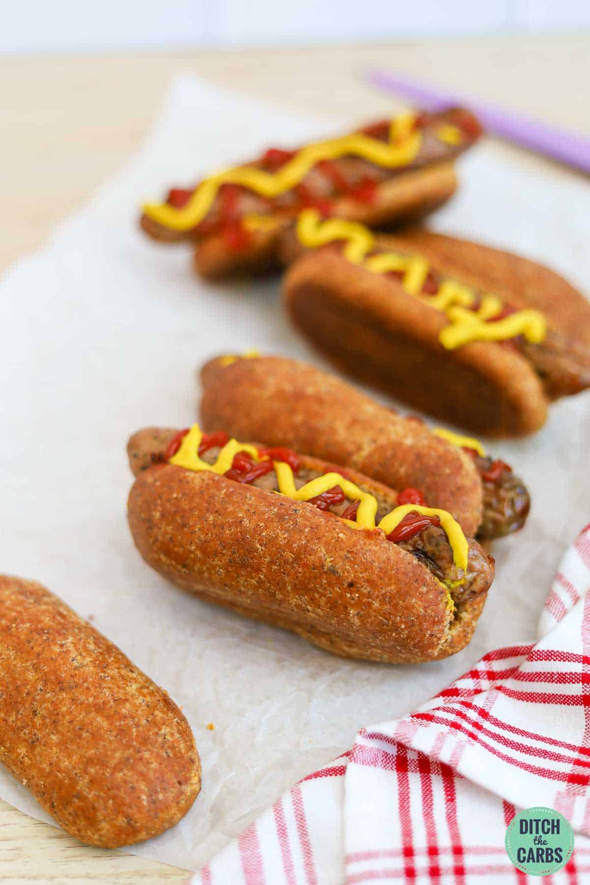 Four keto hot dog buns stuffed with hot dogs and topped with yellow mustard and sugar-free ketchup on a piece of parchment paper.